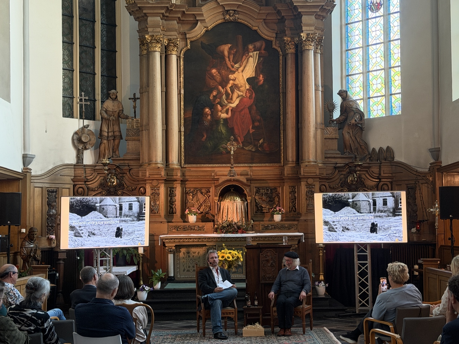 Een gesprek voor het publiek tijdens de jubileumviering, met historische foto's op twee schermen.