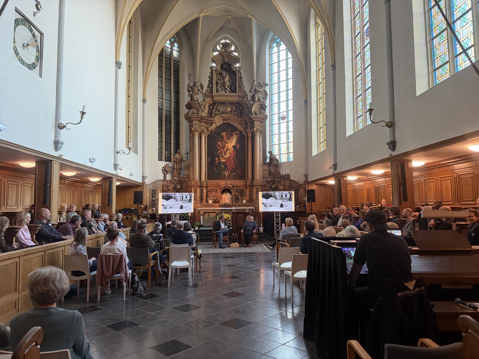 Bezoekers van de jubileumbijeenkomst zitten in de Paterskerk, met vooraan een gesprek en twee presentatieschermen.